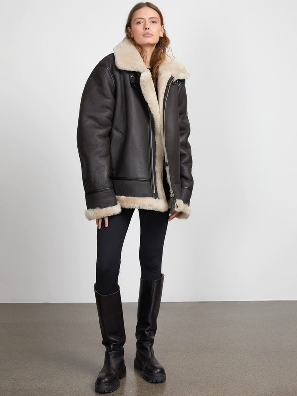 A model showcases a stylish black leather jacket with a plush cream shearling collar, paired with black leggings and knee-high boots. MEOTINE.