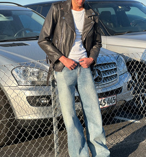 A model wearing a black leather jacket and light blue jeans stands by a fence, showcasing MEOTINE's stylish urban look.
