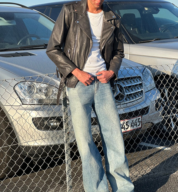 A model wearing a black leather jacket and light blue jeans stands by a fence, showcasing MEOTINE's stylish urban look.