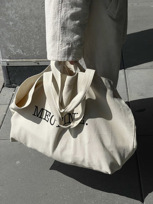 A person holds a large beige tote bag with "MEOTINE." printed on it, showcasing a stylish and practical accessory.