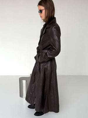 A model showcases a stylish brown leather trench coat by MEOTINE, featuring a belted waist and sleek design, set against a minimalist backdrop.