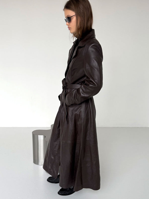 A model showcases a stylish brown leather trench coat by MEOTINE, featuring a belted waist and sleek design, set against a minimalist backdrop.
