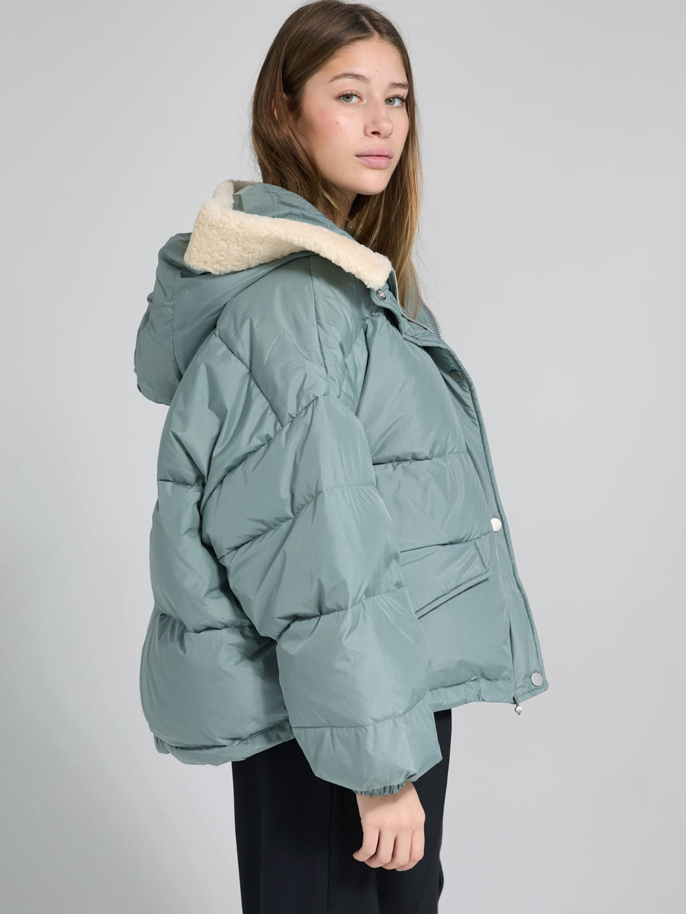 A model wears a cozy, oversized teal puffer jacket with a fur-lined hood, paired with black wide-leg pants. MEOTINE style.