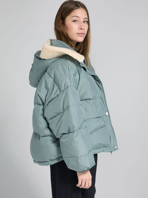 A model showcases a cozy, oversized teal puffer jacket with a soft cream hood, perfect for winter, by MEOTINE.