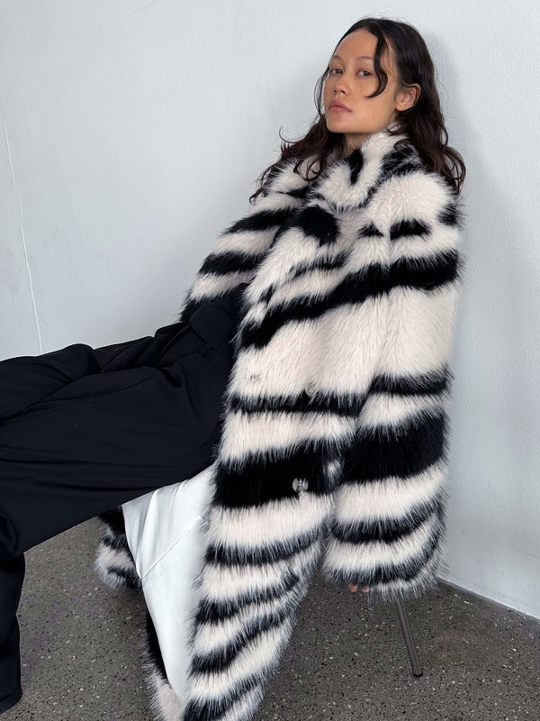 A model poses in a stylish black and white striped faux fur coat by MEOTINE, paired with chic black trousers against a minimalist backdrop.