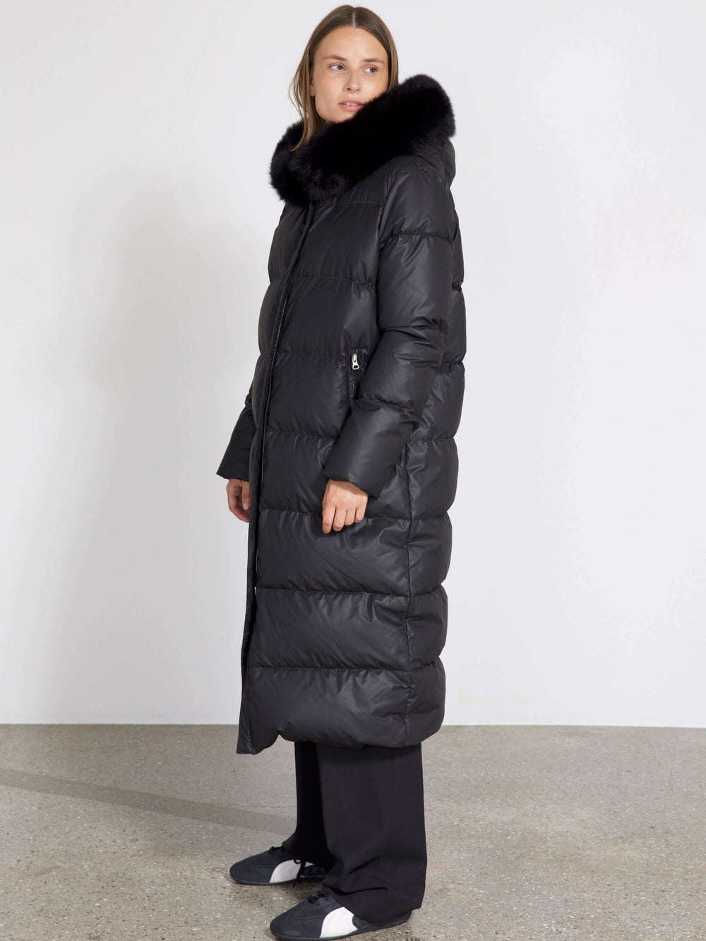 Long black puffer coat with a luxurious fur collar, worn by a model standing against a plain background. MEOTINE brand style.