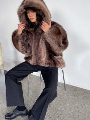 Cozy brown faux fur jacket with a hood, paired with black trousers, worn by a model sitting on a chair. MEOTINE style.