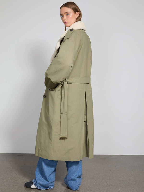 A model wearing a long olive green coat with a fur collar, styled with loose blue jeans, showcasing MEOTINE's chic design.