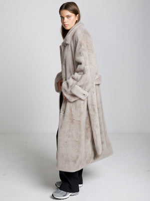 A model wearing a soft, oversized beige faux fur coat by MEOTINE, styled with black pants and sneakers against a neutral backdrop.