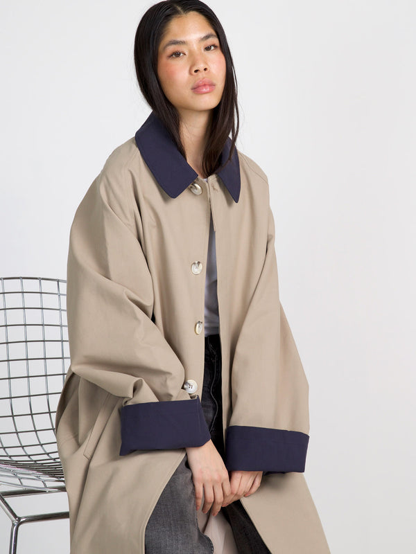 A model wearing a stylish beige MEOTINE coat with a navy collar and cuffs, sitting on a modern wire chair.