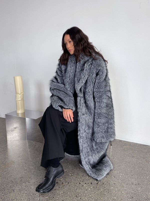 A model sits elegantly in a plush gray MEOTINE fur coat, paired with black attire, showcasing a chic winter look.