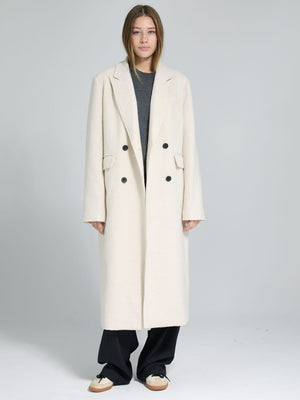 A model wearing a long, cream-colored double-breasted coat by MEOTINE, paired with a gray top and black pants.
