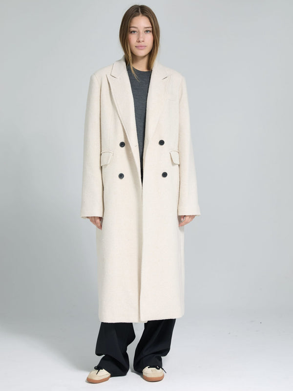 A model wearing a long, cream-colored double-breasted coat by MEOTINE, paired with a gray top and black pants.