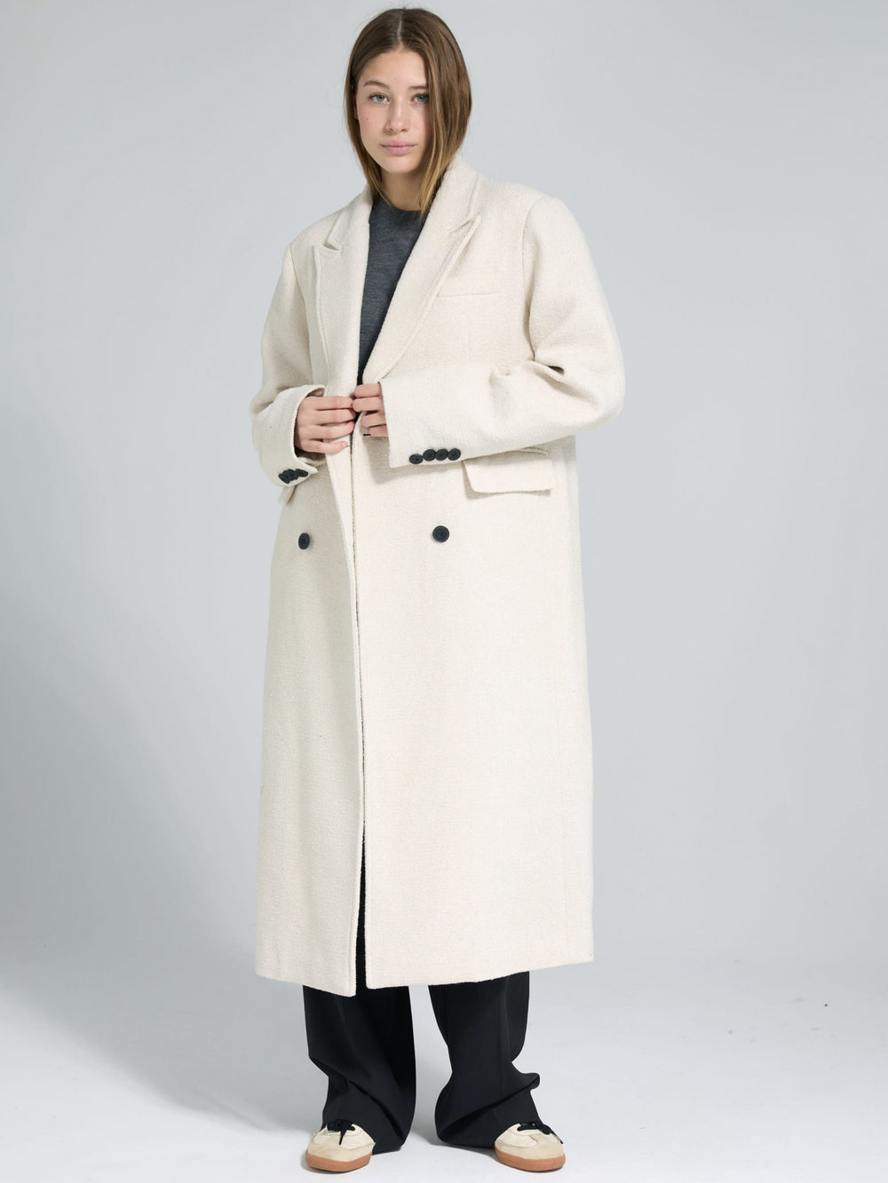 A model wearing a long, cream-colored double-breasted coat by MEOTINE, paired with a gray top and black pants.