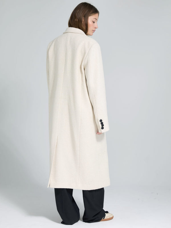 A woman in a long, cream-colored coat with a tailored fit, featuring a back vent and buttoned cuffs, styled with black pants.