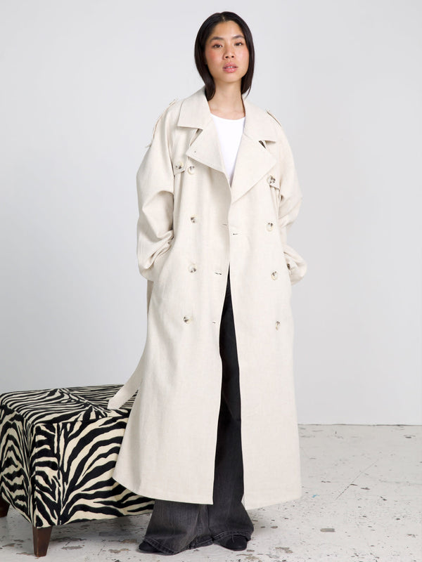 A model wears a stylish beige trench coat by MEOTINE, paired with a simple white top and dark pants, set against a minimalist background.