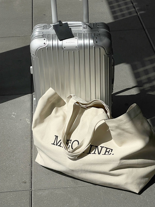 A silver suitcase beside a large beige tote bag with "MEOTINE." The scene is set on a modern, sunlit floor.