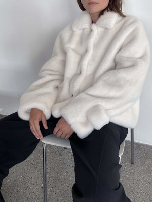 Cozy white faux fur jacket with a button-up front, worn by a model sitting on a chair, showcasing MEOTINE's stylish design.