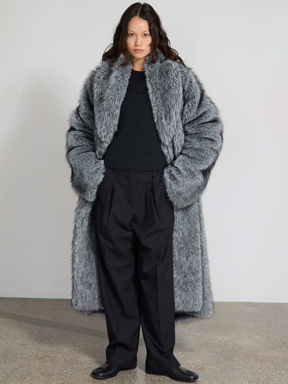 A model wearing a luxurious gray faux fur coat by MEOTINE, styled with black pants, poses against a minimalist backdrop.