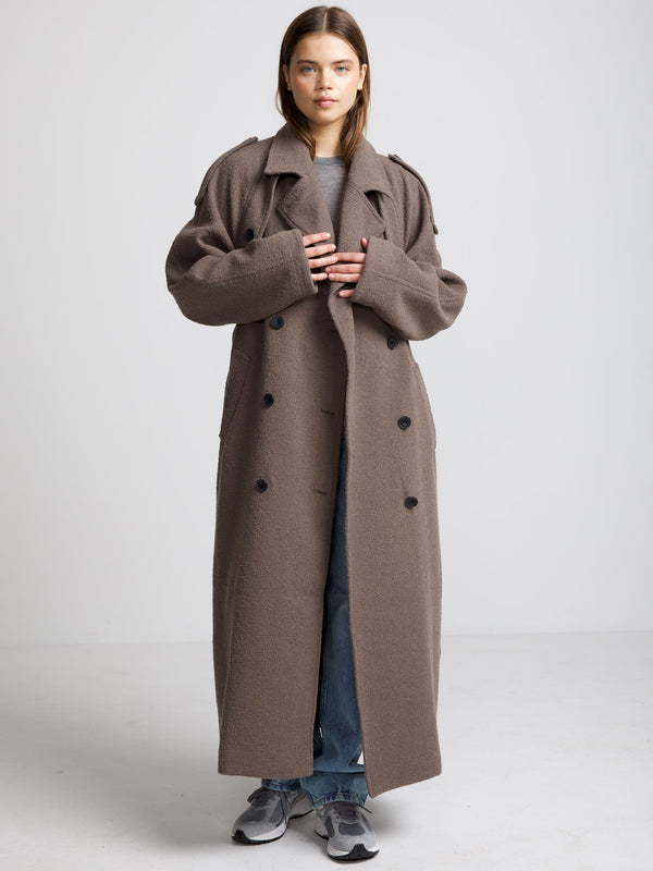 A model wears a long, oversized brown coat with a double-breasted design, styled with casual jeans and sneakers by MEOTINE.