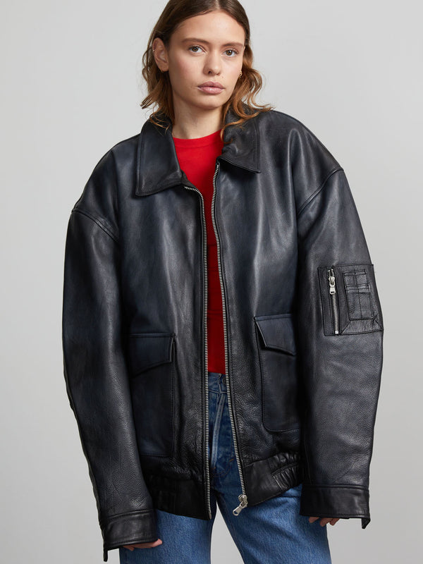 A model wears an oversized black leather jacket with a zip closure and multiple pockets, styled with a red top underneath.