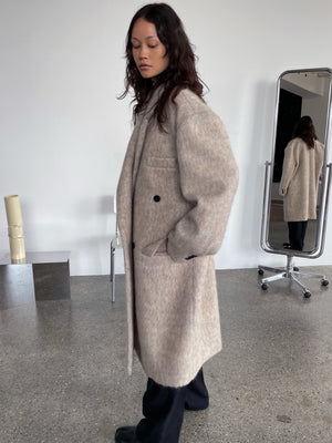 A model wearing a cozy beige MEOTINE coat, featuring a relaxed fit and double-breasted design, stands in a minimalist setting.