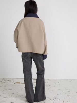 A model wearing a beige oversized jacket with a navy collar, paired with flared gray jeans, showcasing a modern, casual style.
