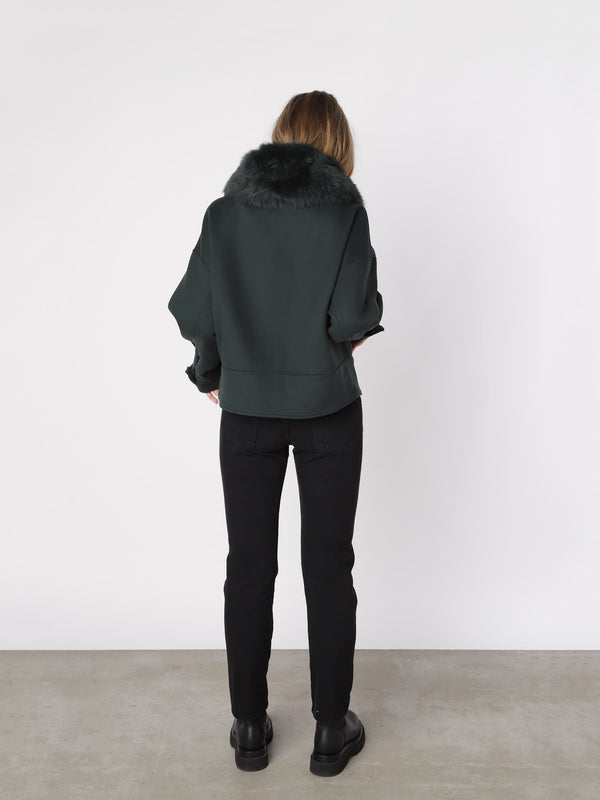A woman stands with her back to the camera, wearing a dark green MEOTINE jacket with a fur collar, paired with black jeans.