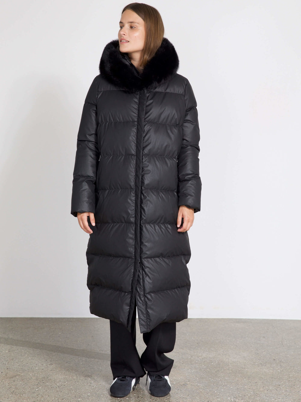 Long black puffer coat with a luxurious fur collar, worn by a model standing against a plain background. MEOTINE brand style.