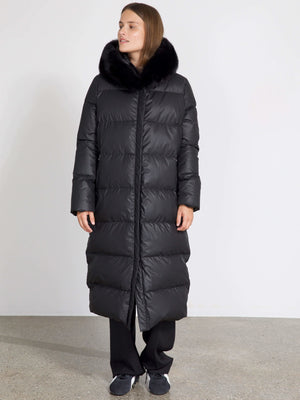 Long black puffer coat with a luxurious fur collar, worn by a model standing against a plain background. MEOTINE brand style.