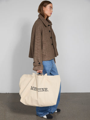 A model wears a brown textured jacket with a large MEOTINE tote bag, paired with blue jeans and black shoes, against a neutral backdrop.