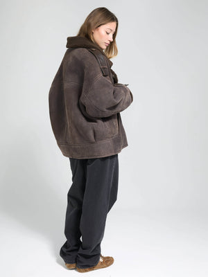 A model wearing a brown oversized MEOTINE jacket with a cozy collar, paired with loose black pants, standing against a light background.