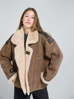Brown shearling jacket with a plush collar and leather accents, worn by a model against a light gray background. MEOTINE brand.