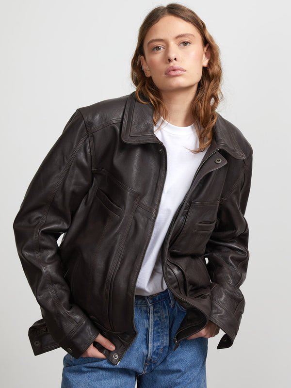 A model wears a stylish brown leather jacket from MEOTINE, paired with a simple white t-shirt and relaxed blue jeans.