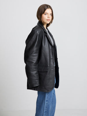 A model showcases a stylish black leather blazer from MEOTINE, paired with a casual gray top and blue jeans.